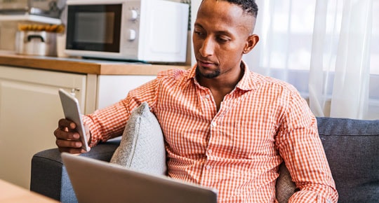 A man sits on a sofa in front of an open laptop while scrolling on his mobile phone.